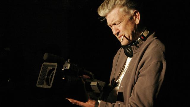 David Lynch hält konzentriert eine laufende Kamera in beiden Händen, eine Zigarette im Mundwinkel und Kopfhörer um den Hals