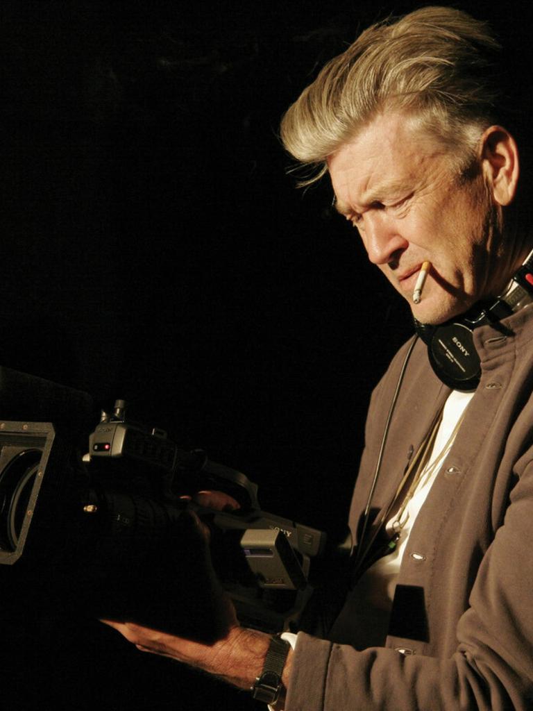 David Lynch hält konzentriert eine laufende Kamera in beiden Händen, eine Zigarette im Mundwinkel und Kopfhörer um den Hals