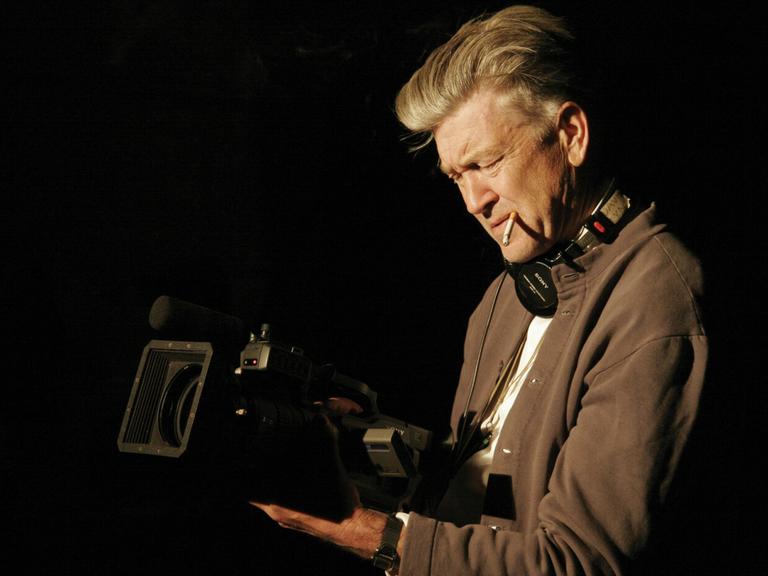 David Lynch hält konzentriert eine laufende Kamera in beiden Händen, eine Zigarette im Mundwinkel und Kopfhörer um den Hals