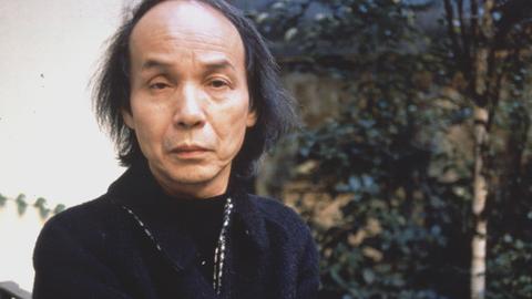 Toru Takemitsu blickt auf einem Foto von 1989 ernst in die Kamera, während er auf einem Gartenstuhl sitzt.