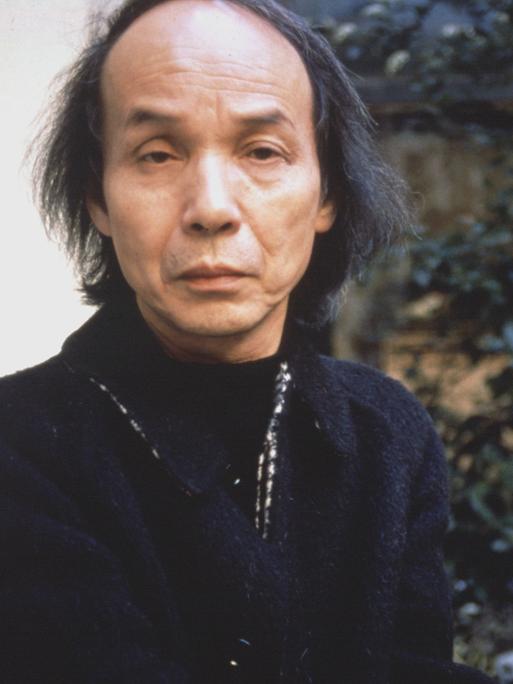 Toru Takemitsu blickt auf einem Foto von 1989 ernst in die Kamera, während er auf einem Gartenstuhl sitzt.