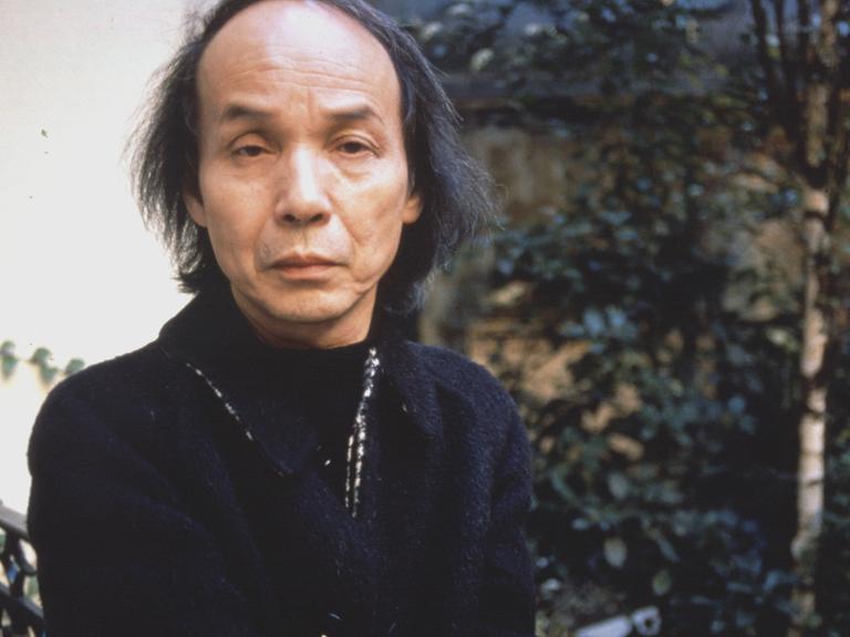 Toru Takemitsu blickt auf einem Foto von 1989 ernst in die Kamera, während er auf einem Gartenstuhl sitzt.