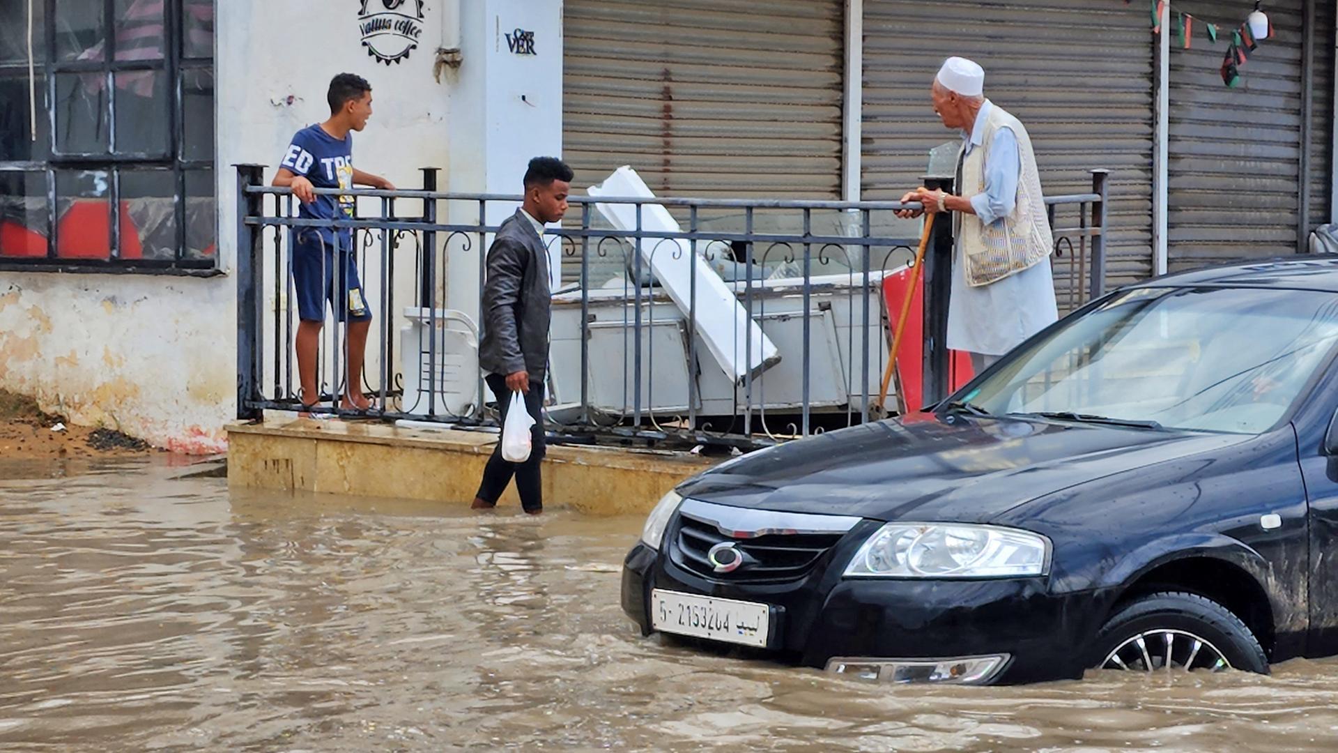 Ein Auto fährt durch das kniehohe Wasser, ein Mann watet hindurch. Auf einer Terasse vor einem Gebäude dahinter stehen ein Jugendlicher und ein älterer Mann.