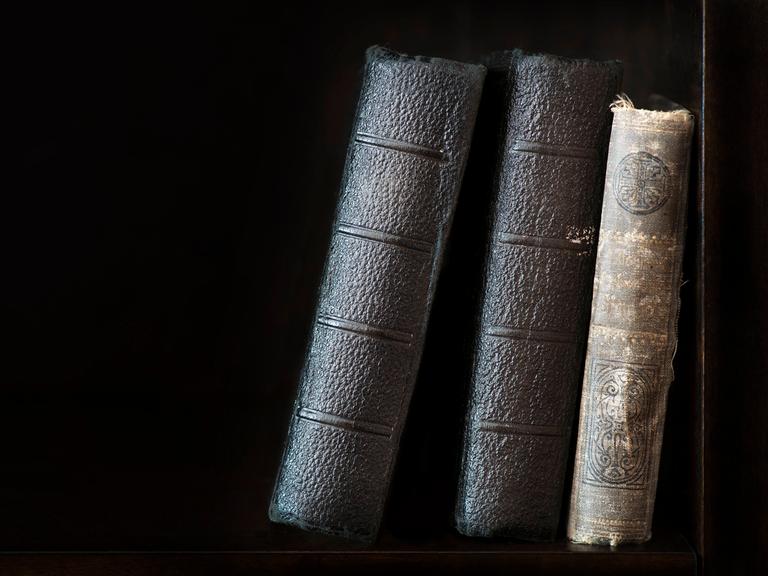 Drei alte, historische Bücher stehen aneinander gelehnt in einem Regal