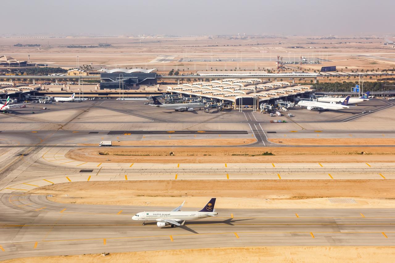 Man sieht im Bildvordergrund ein Flugzeug auf der Start- oder Landebahn. Dahinter Flughafengebäude und weitere geparkte Maschinen. 