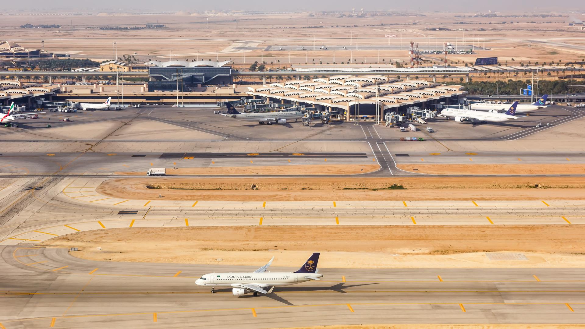 Man sieht im Bildvordergrund ein Flugzeug auf der Start- oder Landebahn. Dahinter Flughafengebäude und weitere geparkte Maschinen. 