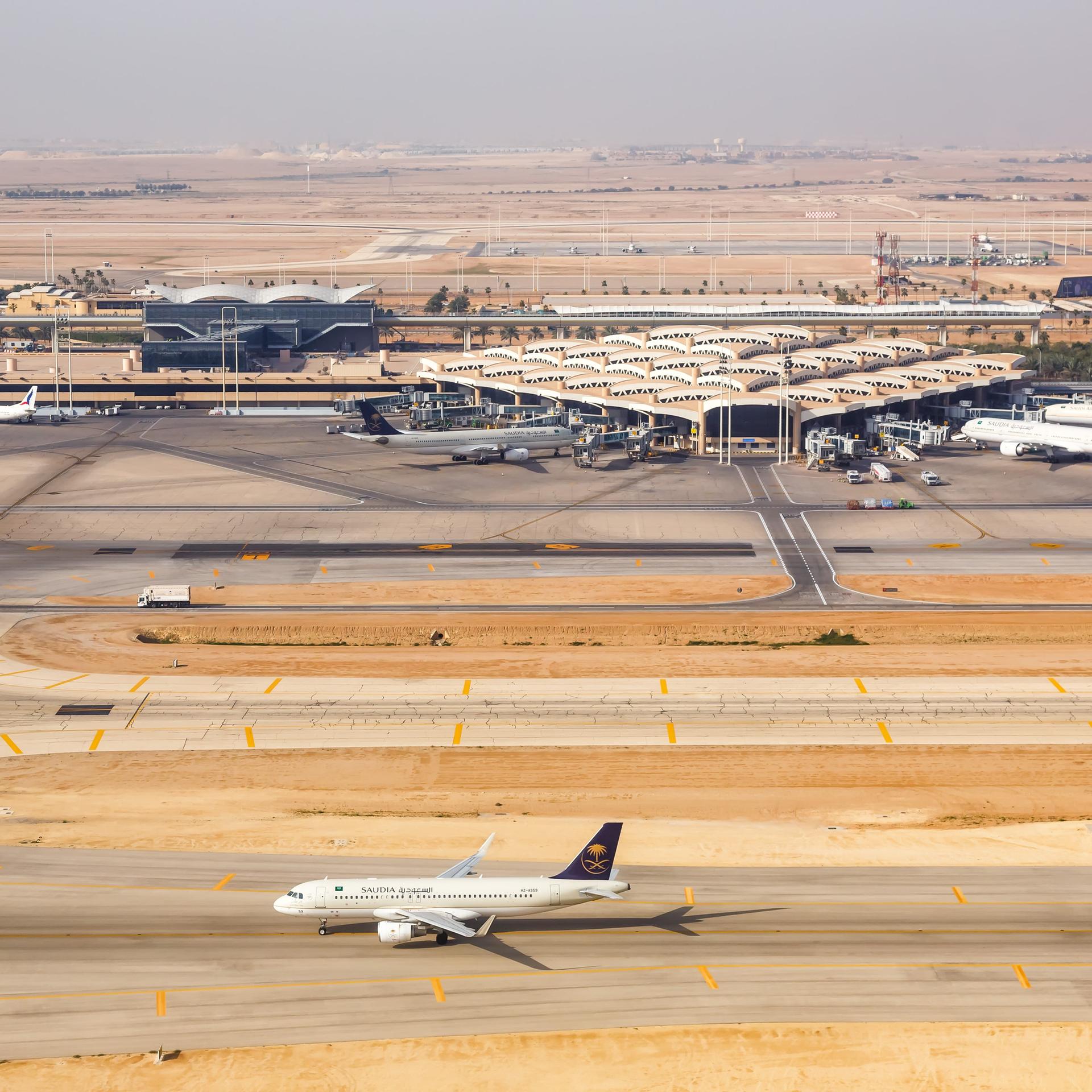 Man sieht im Bildvordergrund ein Flugzeug auf der Start- oder Landebahn. Dahinter Flughafengebäude und weitere geparkte Maschinen. 