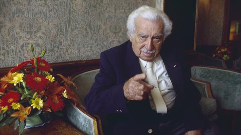 Jorge Amado sitzt in einer Lobby auf einem Sessel und deutet mit ernstem Blick in die Kamera.