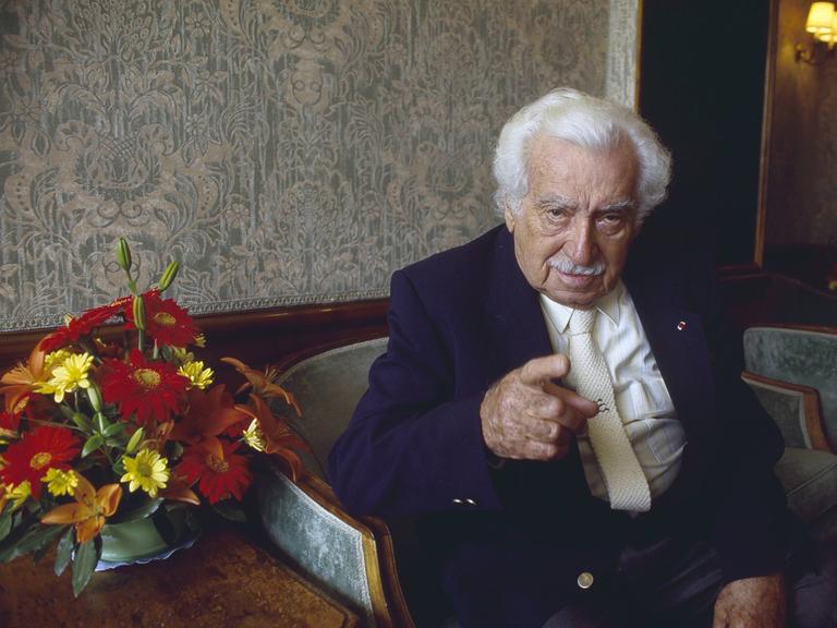 Jorge Amado sitzt in einer Lobby auf einem Sessel und deutet mit ernstem Blick in die Kamera. Jorge Amado sitzt in einer Lobby auf einem Sessel und deutet mit ernstem Blick in die Kamera.