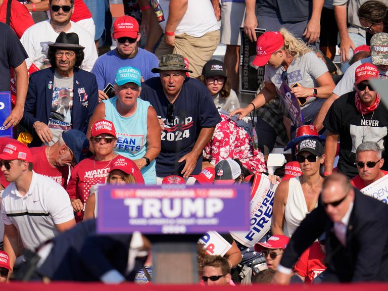 Das Rednerpult von Donald Trump bei einer Wahlkampfveranstaltung in Pennsylvania. Der Ex-Präsident duckt sich (nicht sichtbar) dahinter, ein Bodyguard kommt von recht, die Zuschauer schauen erschrocken. 