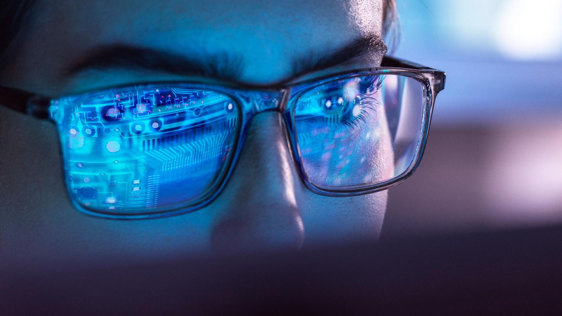Nahaufnahme eines Mannes in dessen Brillengäsern sich Datenleitungen bzw. ein Computerchip spiegeln.