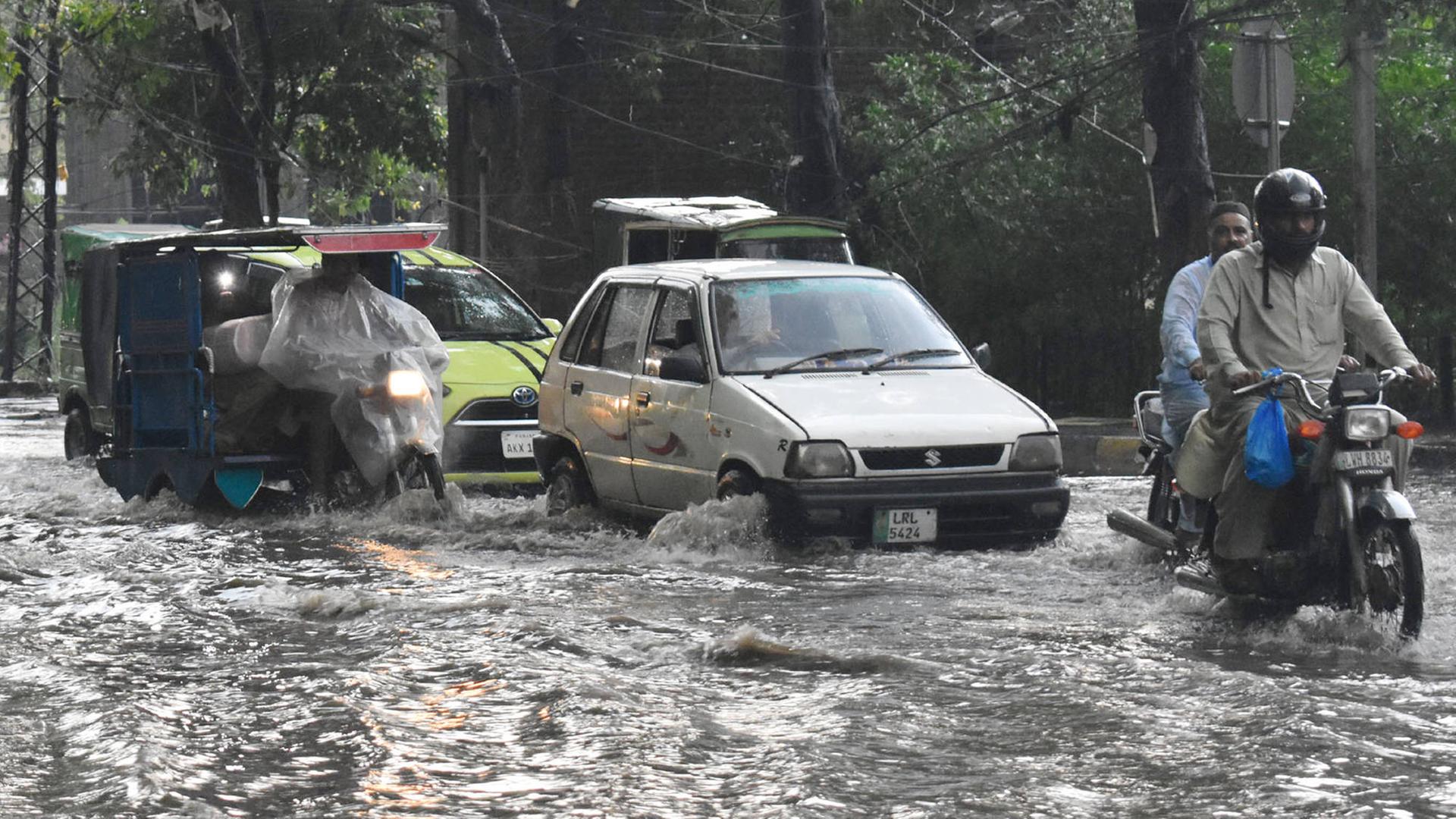 Zwei Autos und drei Krafträder fahren auf einer Straße, auf der das Wasser zentimeterhoch steht. Dahinter Bäume und Gebäude