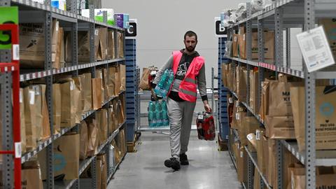 Automatisiertes Distributionszentrum des tschechischen Online-Lebensmittelhändlers Rohlik in Chrastany bei Prag. Ein Mitarbeiter in Warnweste läuft mit Lebensmitteln durch Regalreihen mit vollgepackten Paketen und Taschen