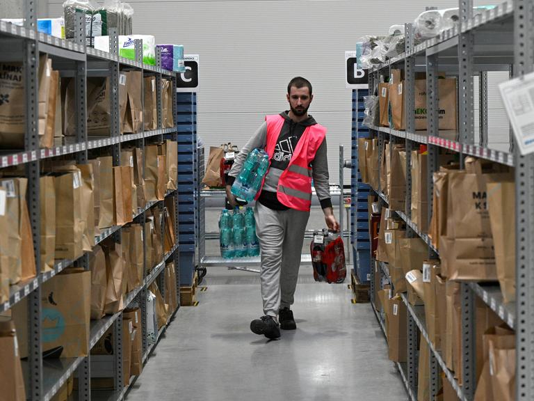 Automatisiertes Distributionszentrum des tschechischen Online-Lebensmittelhändlers Rohlik in Chrastany bei Prag. Ein Mitarbeiter in Warnweste läuft mit Lebensmitteln durch Regalreihen mit vollgepackten Paketen und Taschen