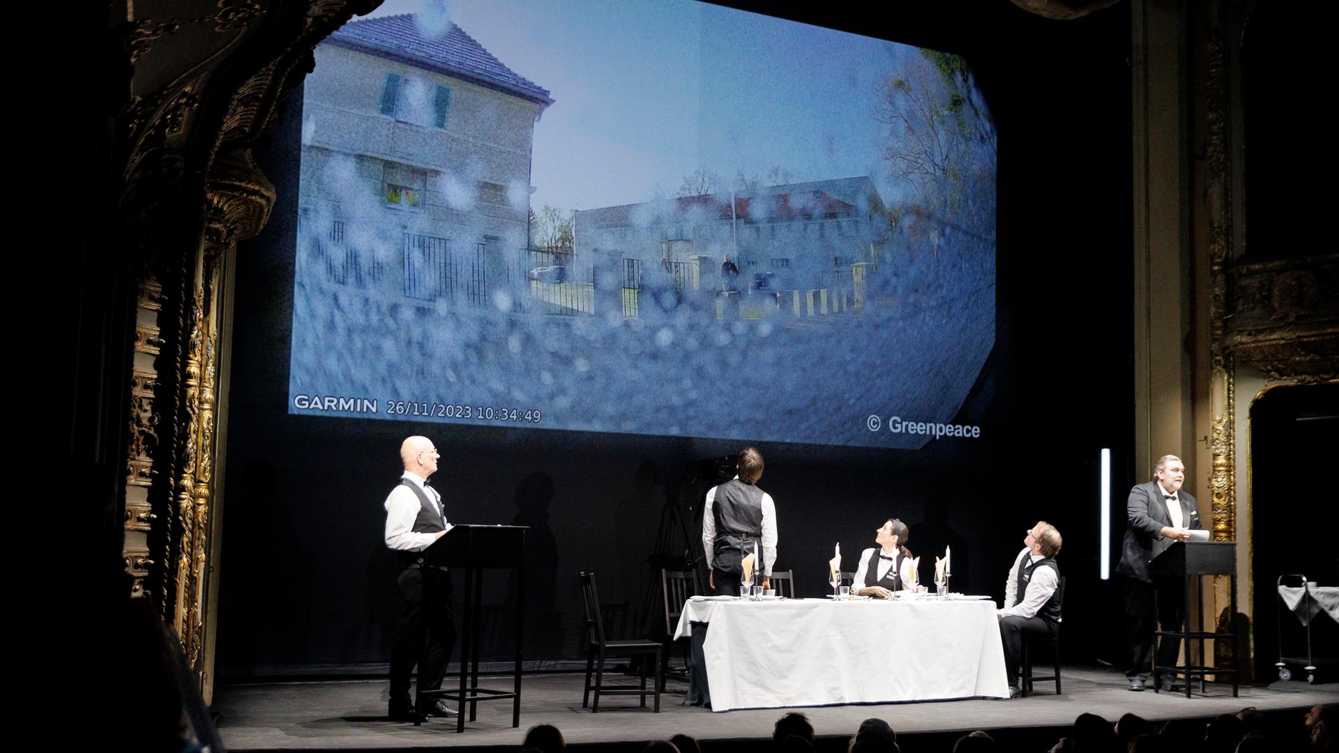 Fünf Schauspieler stehen und sitzen auf der Bühne des Berliner Ensembles an zwei Pulten und um einen Tisch, im Hintergrund wird ein Fernsehbild projiziert.