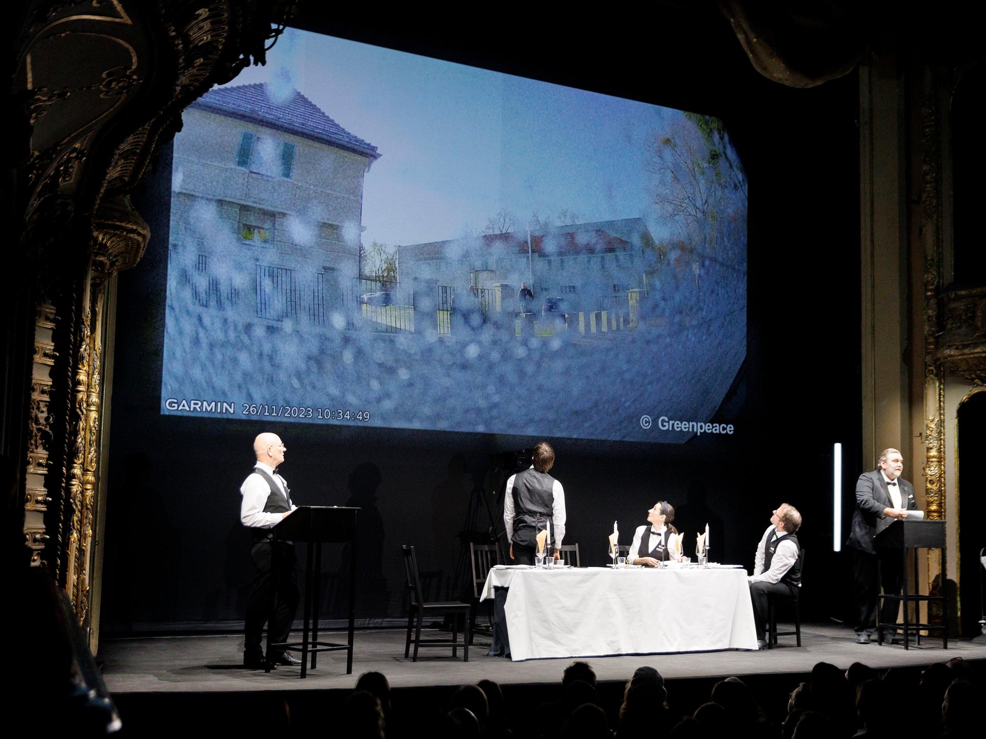 Fünf Schauspieler stehen und sitzen auf der Bühne des Berliner Ensembles an zwei Pulten und um einen Tisch, im Hintergrund wird ein Fernsehbild projiziert.
