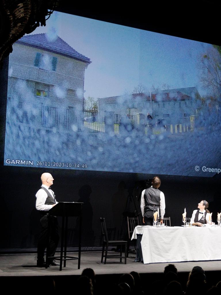 Fünf Schauspieler stehen und sitzen auf der Bühne des Berliner Ensembles an zwei Pulten und um einen Tisch, im Hintergrund wird ein Fernsehbild projiziert.