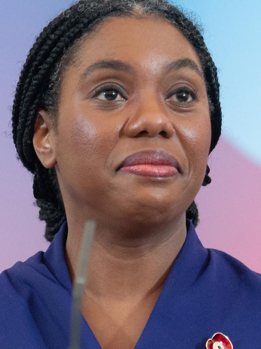 Ein Porträt von Kemi Badenoch. Sie steht hinter zwei Mikronen und lächelt mit heruntergezogenen Mundwinkeln. Badenoch ist schwarz. Ihre langen Haare sind hinter dem Kopf zusammengebunden. 