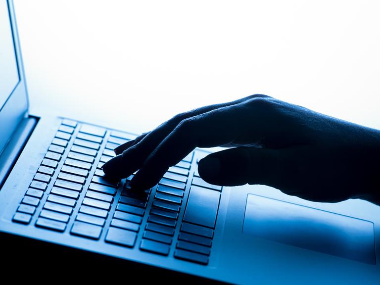Eine Frauenhand in Nahaufnahme tippt auf einem Laptop, die Szene wirkt durch blaues Licht kühl und technisch.