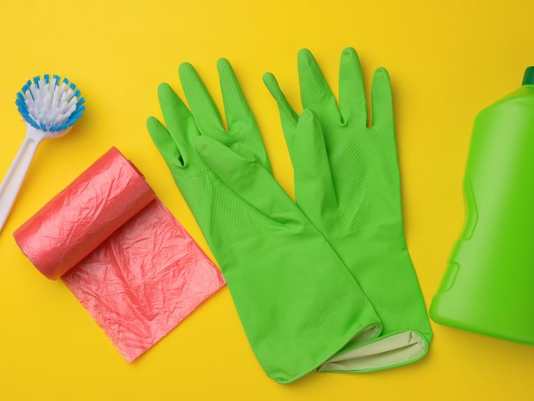 Grüne Gummihandschuhe für die Reinigung, roter Mülleimer, Plastiktütenrolle und Plastikflasche mit Reinigungsmittel auf gelbem Hintergrund (Draufsicht)