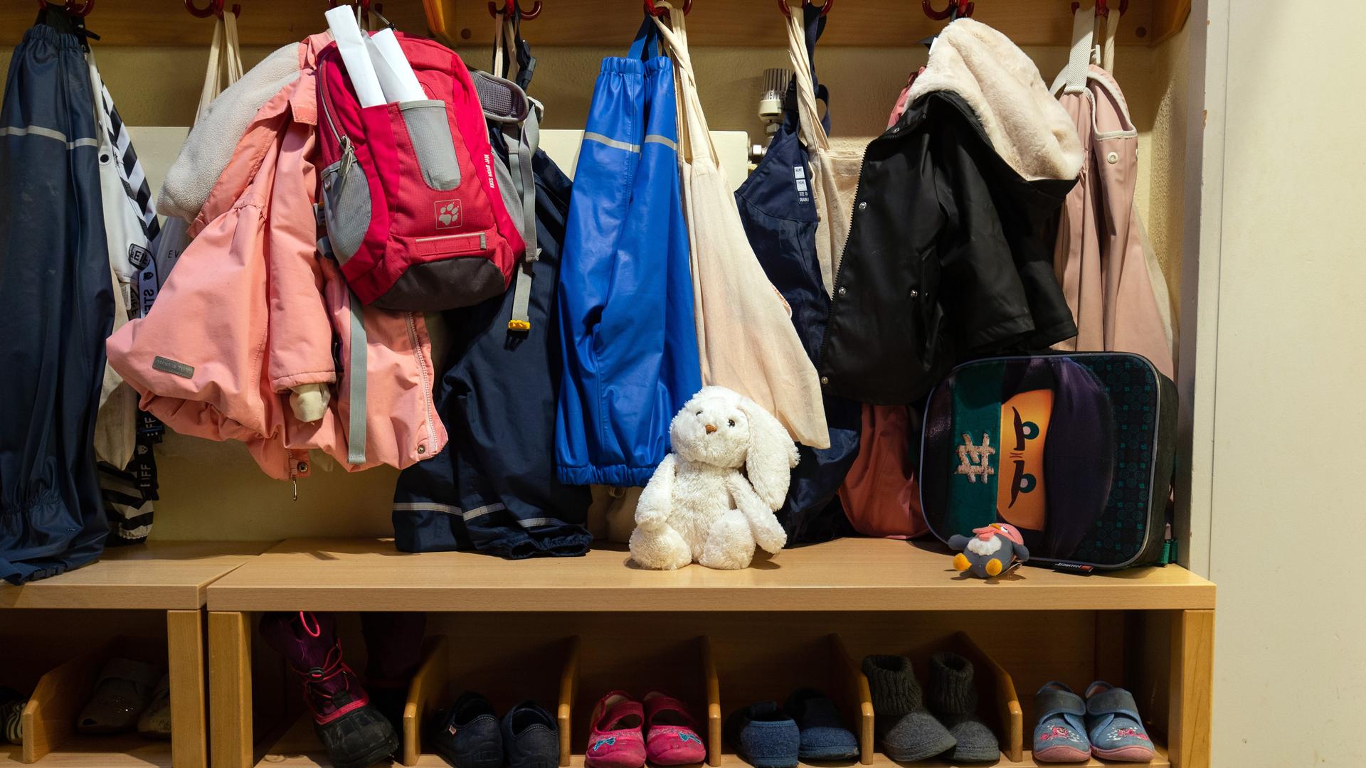 In der Garderobe einer Kita hängen Kinderjacken und -rucksäcke, darunter stehen Kinderschuhe. In der Mitte sitzt ein weißer Stoffhase. 