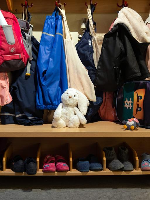 In der Garderobe einer Kita hängen Kinderjacken und -rucksäcke, darunter stehen Kinderschuhe. In der Mitte sitzt ein weißer Stoffhase. 