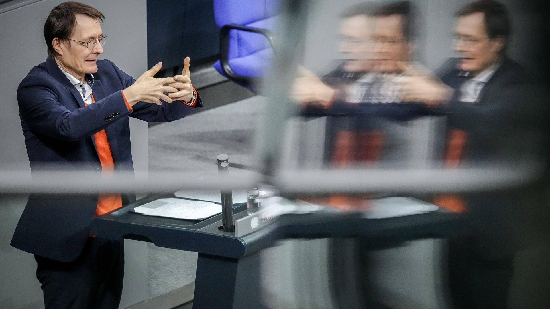 Lauterbach steht am Rednerpult und gestikuliert mit beiden Händen. Er spiegelt sich in einer Scheibe.