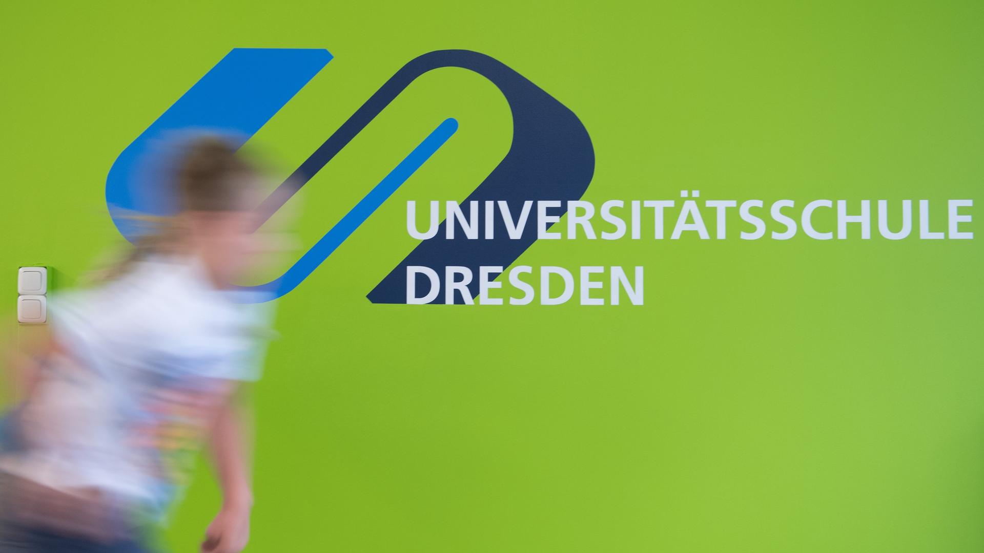 Zu sehen ist ein Mädchen, unscharf, das an dem Logo der Universitätsschule Dresden vorbei geht.