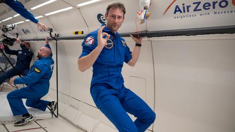 Der polnische Astronaut Sławosz Uznański in einem plauen ESA-Overall macht während eines Parabelflugs in der Schwerelosigkeit mit der Hand ein Okay-Zeichen