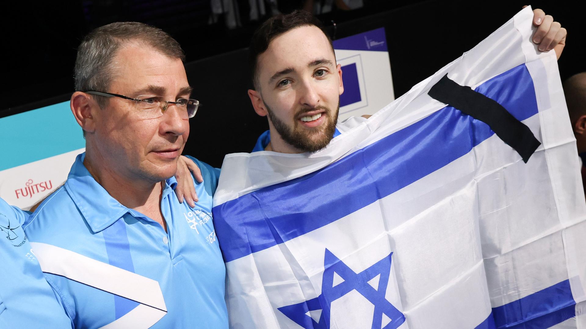 Turn-WM 2023 in Antwerpen: Ein schwarzes Trauerband ist auf der israelischen Flagge zu sehen, die der Turner Artem Dolgopyat hochhält, während er und sein Trainer den Gewinn einer Goldmedaille feiern. Turn-WM 2023 in Antwerpen: Ein schwarzes Trauerband ist auf der israelischen Flagge zu sehen, die der Turner Artem Dolgopyat hochhält, während er und sein Trainer den Gewinn einer Goldmedaille feiern.