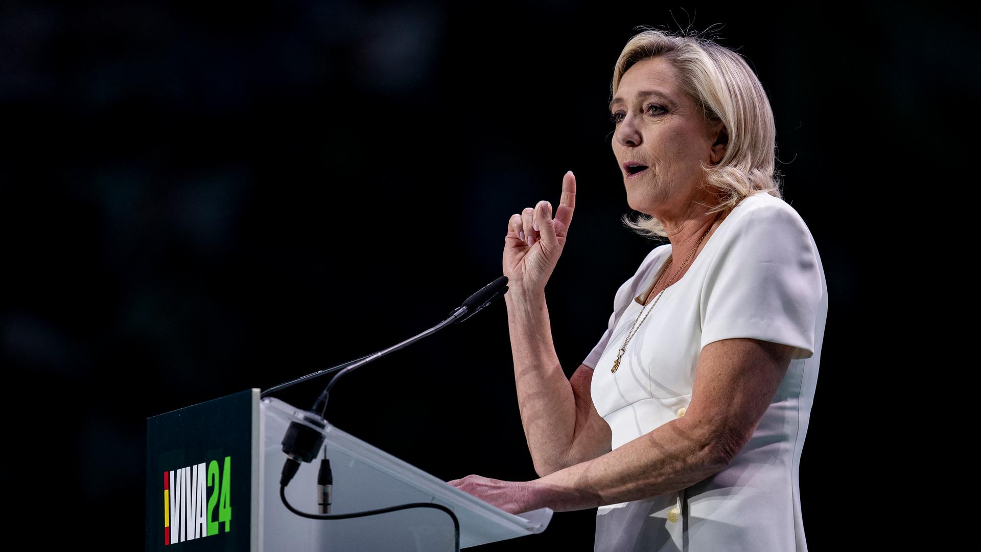 Le Pen im weißen Sommerkleid steht an einem Rednepult, spricht und hebt dabei den rechten Zeigefinger. Der Hintergrund ist dunkel.