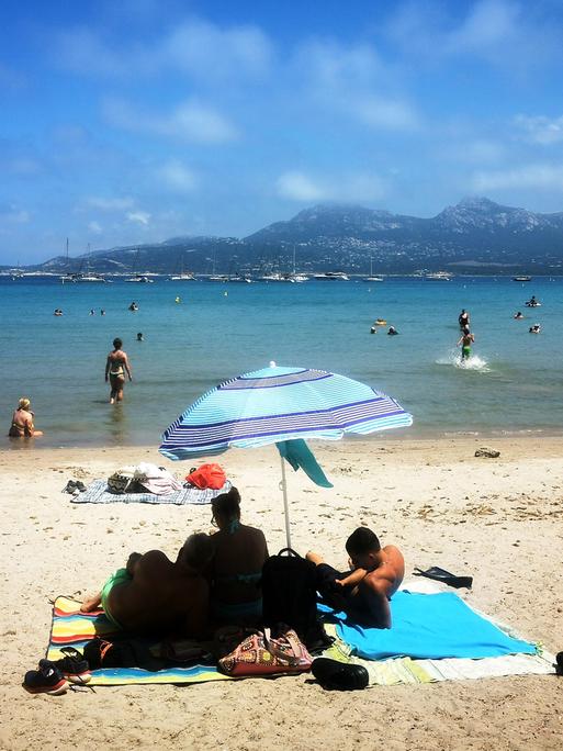 Menschen liegen am Strand von Korsika unter Sonnenschirmen. 