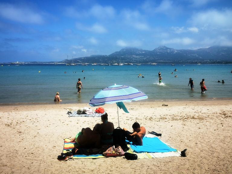 Menschen liegen am Strand von Korsika unter Sonnenschirmen. 