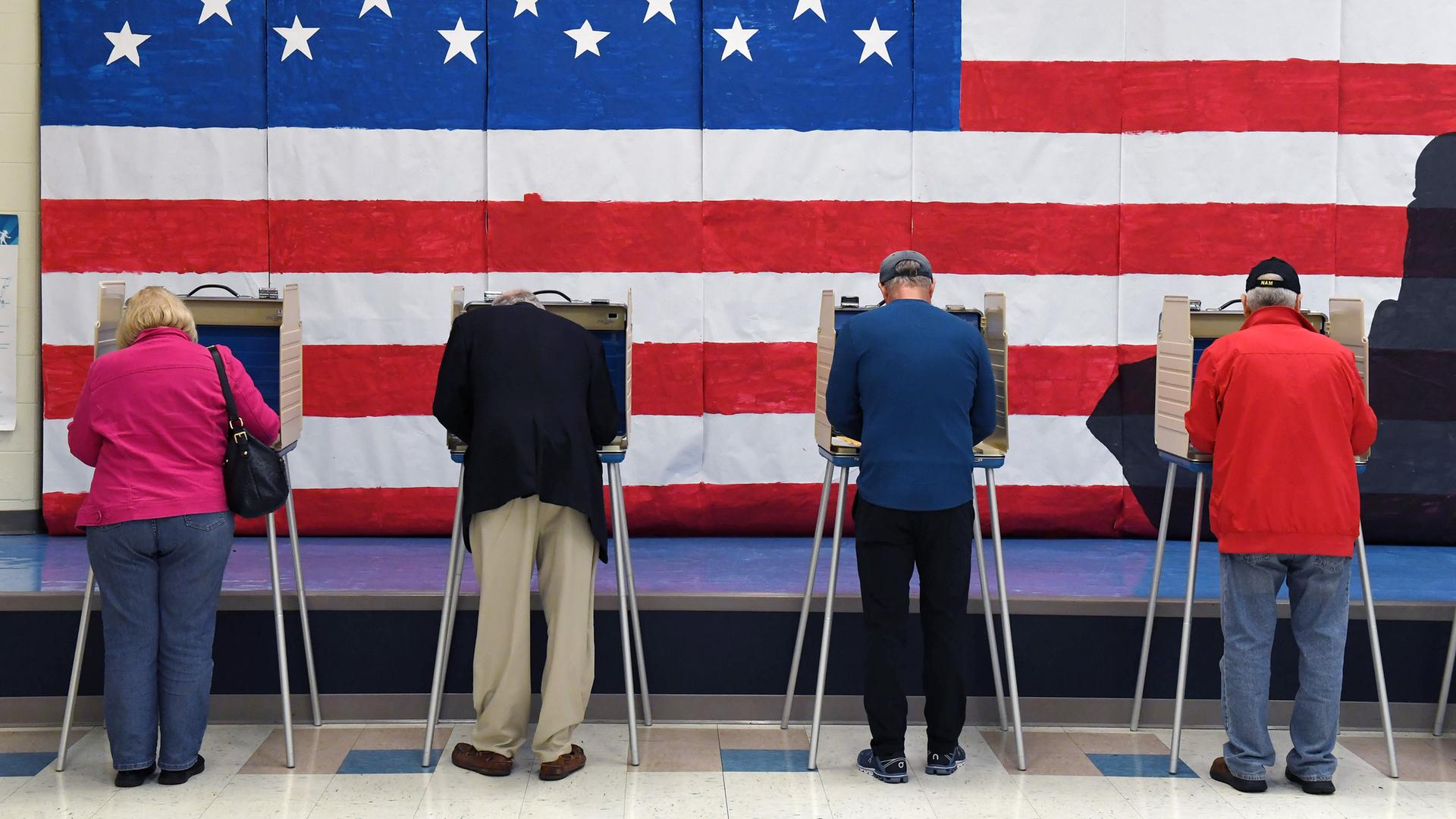 Mehrere Personen stehen an Wahlurnen, an der Wand dahinter hängt eine große US-Flagge.