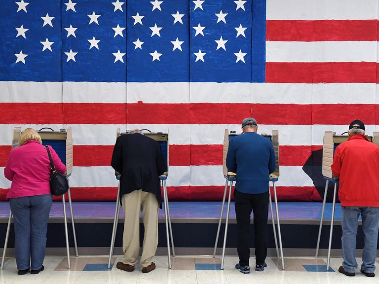 Mehrere Personen stehen an Wahlurnen, an der Wand dahinter hängt eine große US-Flagge.