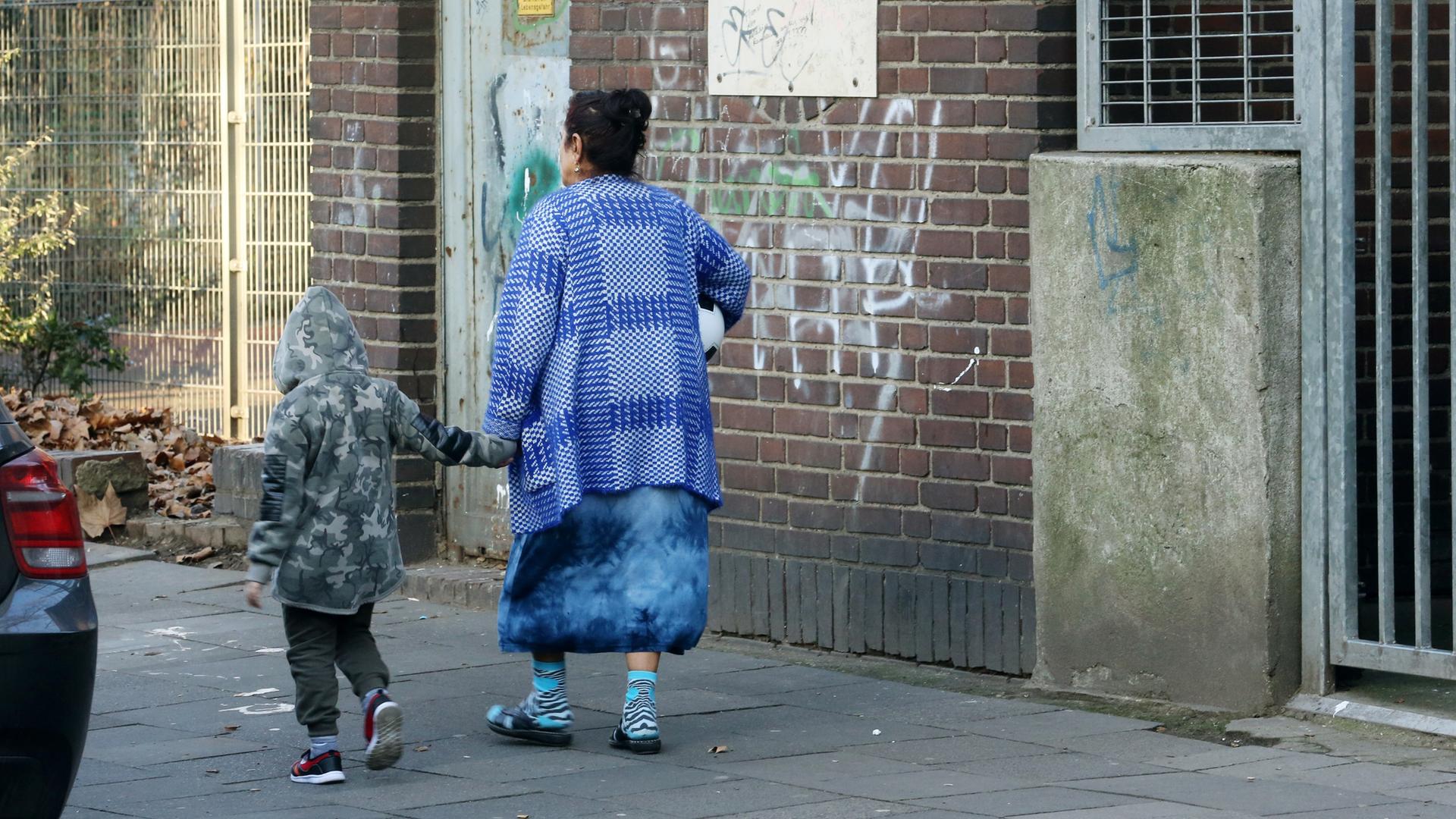 Eine Frau und ein Kind gehen an einem Gebäude vorbei. Die beiden haben abgetragene Kleidung an. Das Gebäude ist dreckig und mit Graffiti besprüht. 