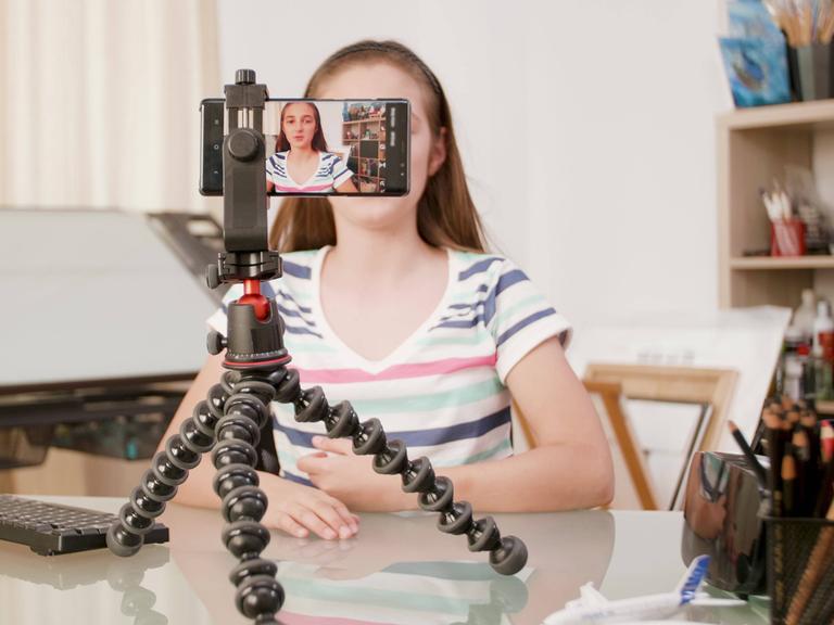 Ein Mädchen sitzt vor einer Handy-Kamera in einem Zimmer und filmt sich selbst.
