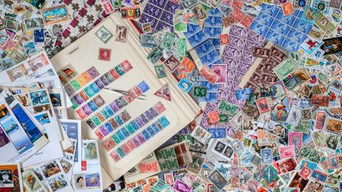 Ein Tisch, auf dem ein Buch mit Briefmarken zu sehen ist. Drumherum liegen hunderte von anderen Briefmarken herum. Ein Tisch, auf dem ein Buch mit Briefmarken zu sehen ist. Drumherum liegen hunderte von anderen Briefmarken herum.