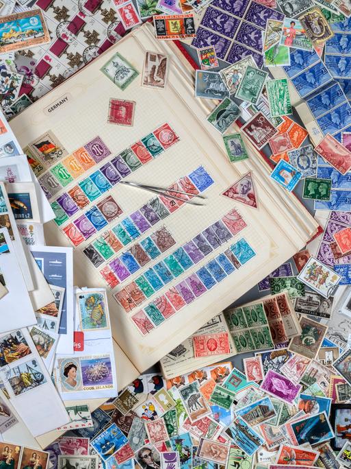 Ein Tisch, auf dem ein Buch mit Briefmarken zu sehen ist. Drumherum liegen hunderte von anderen Briefmarken herum. Ein Tisch, auf dem ein Buch mit Briefmarken zu sehen ist. Drumherum liegen hunderte von anderen Briefmarken herum.