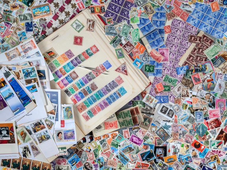 Ein Tisch, auf dem ein Buch mit Briefmarken zu sehen ist. Drumherum liegen hunderte von anderen Briefmarken herum. Ein Tisch, auf dem ein Buch mit Briefmarken zu sehen ist. Drumherum liegen hunderte von anderen Briefmarken herum.