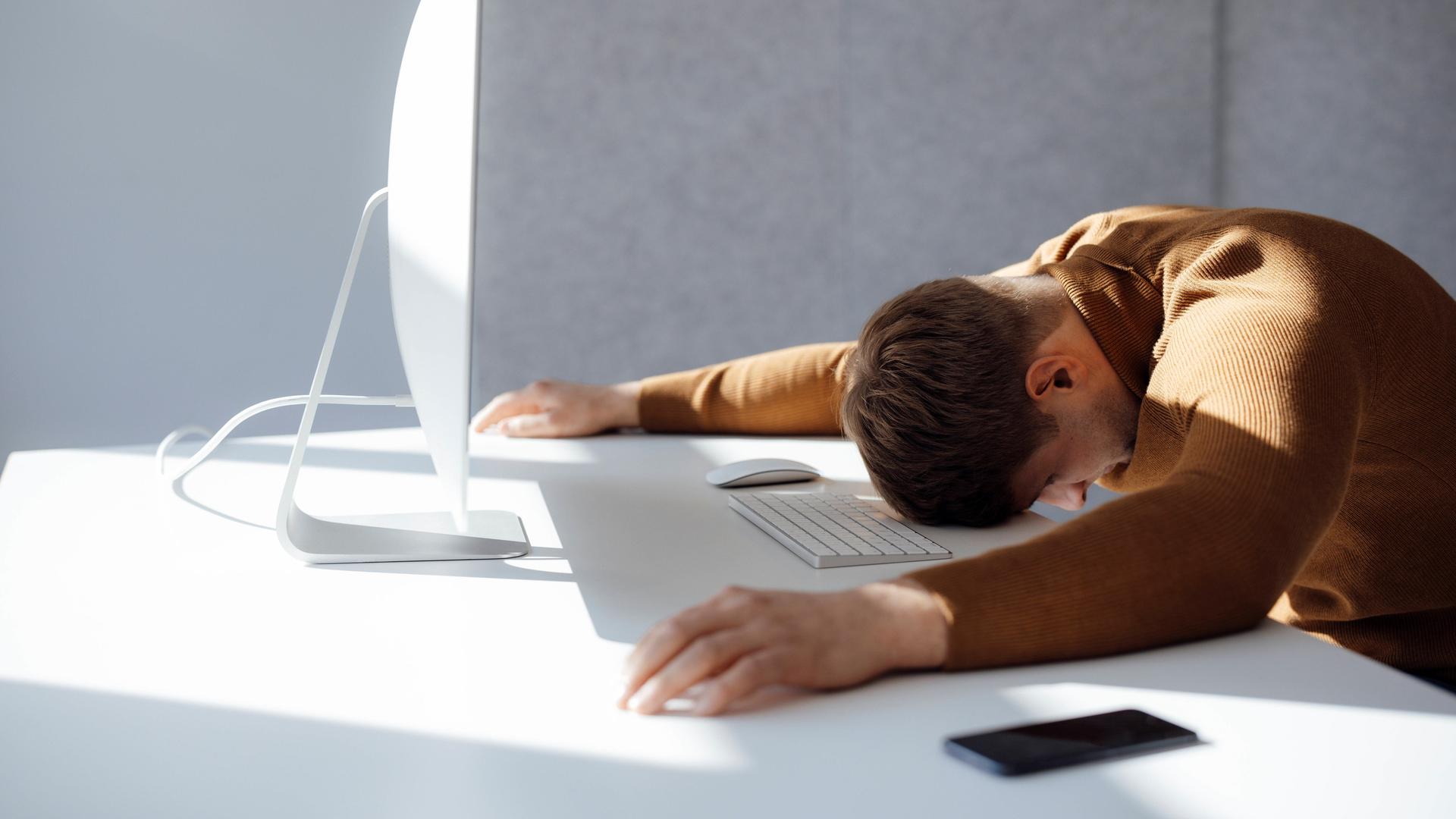 Ein Mann sitzt an einem fast leeren Schreibtisch, er hat den Kopf sowie die Hände und Arme auf die Tischplatte gelegt. Er wirkt erschöpft.