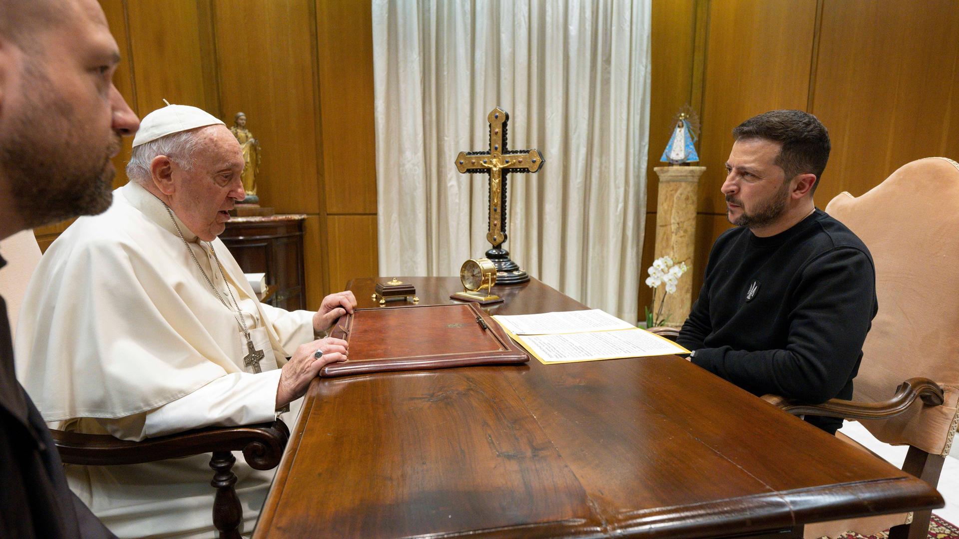 Papst Franziskus sitzt gegenüber dem ukrainischen Präsidenten Wolodymyr Selenskyj an einem dunklen Holztisch. Darauf befindet sich unter anderem ein Kreuz.