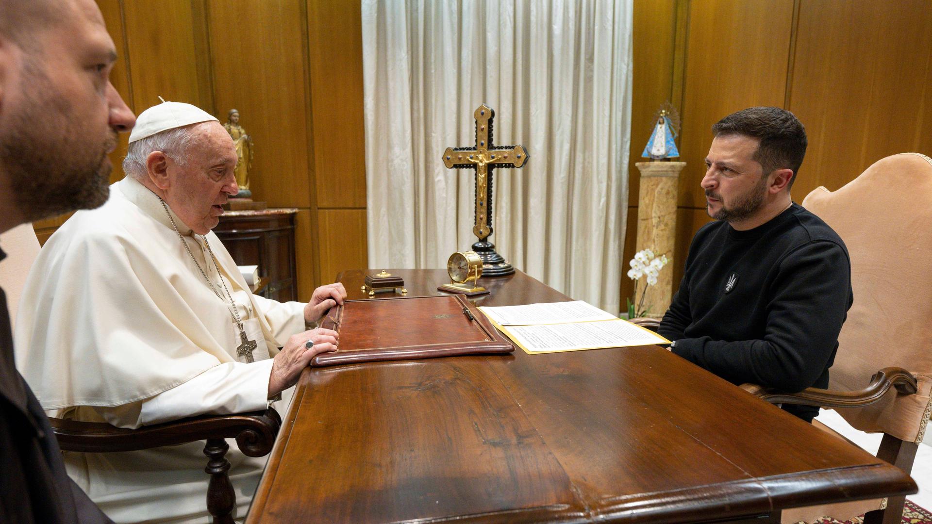 Papst Franziskus sitzt gegenüber dem ukrainischen Präsidenten Wolodymyr Selenskyj an einem dunklen Holztisch. Darauf befindet sich unter anderem ein Kreuz. Papst Franziskus sitzt gegenüber dem ukrainischen Präsidenten Wolodymyr Selenskyj an einem dunklen Holztisch. Darauf befindet sich unter anderem ein Kreuz.