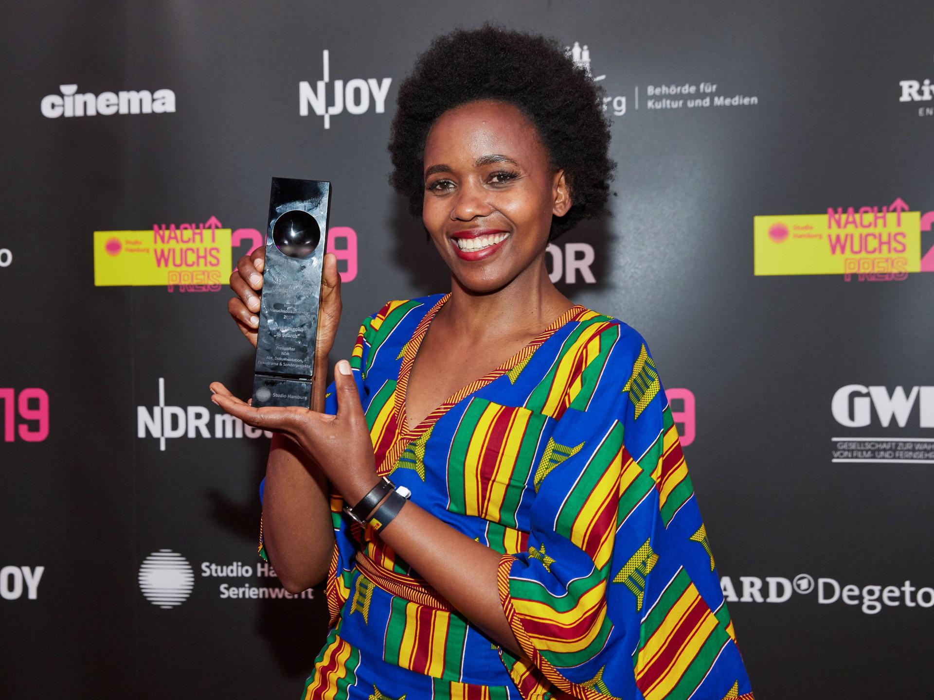 Beryl Magoko hält lächelnd einen Award in die Kamera. Sie trägt ein bunt-gestreiftes Kleid.