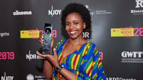 Beryl Magoko hält lächelnd einen Award in die Kamera. Sie trägt ein bunt-gestreiftes Kleid.