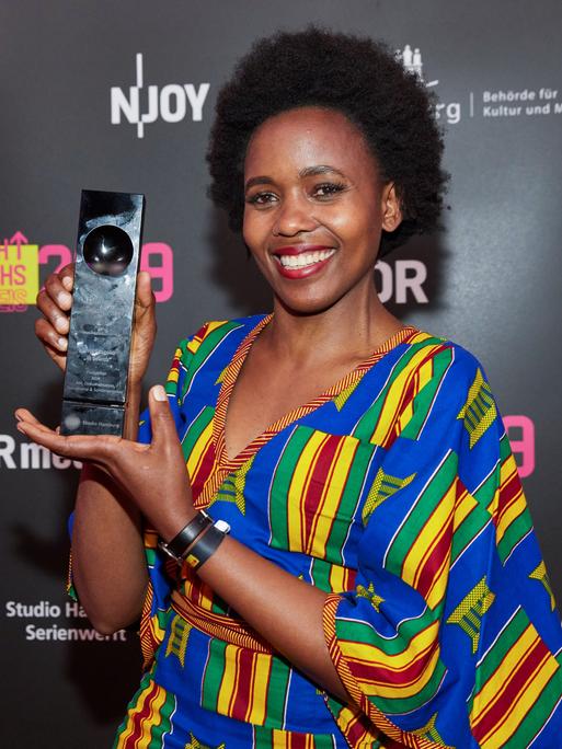 Beryl Magoko hält lächelnd einen Award in die Kamera. Sie trägt ein bunt-gestreiftes Kleid.