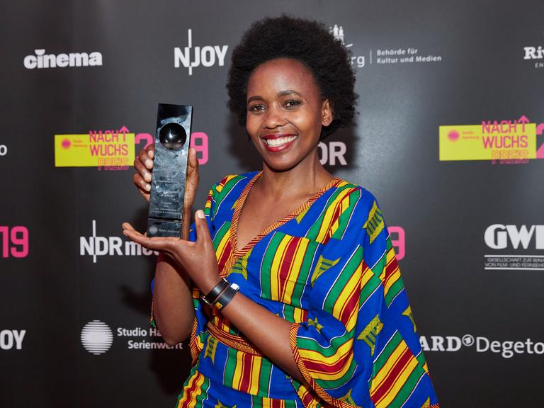 Beryl Magoko hält lächelnd einen Award in die Kamera. Sie trägt ein bunt-gestreiftes Kleid.
