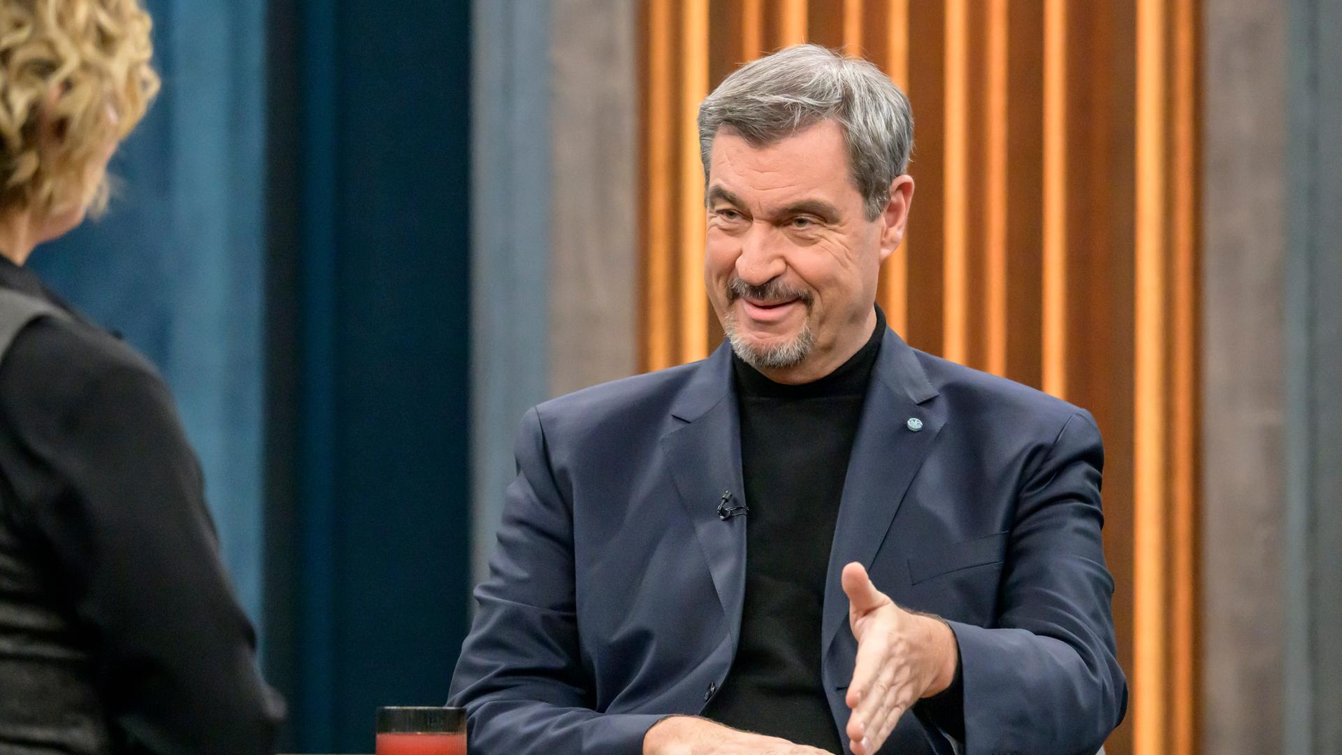 CSU-Chef Söder sitzt an einem Tisch und gestikuliert. Vor ihm in Rückansicht sitzt Caren Miosga.