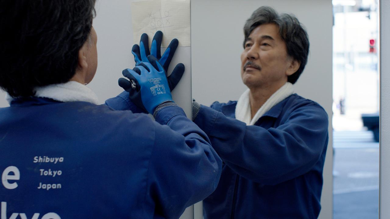 Der Schauspieler Kōji Yakusho steht vor einem Spiegel. Er trägt einen blauen Mantel und blaue Gummihandschuhe.