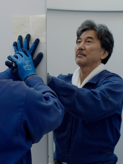 Der Schauspieler Kōji Yakusho steht vor einem Spiegel. Er trägt einen blauen Mantel und blaue Gummihandschuhe.